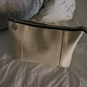 Beige necessär från Necesse - Snygg beige necessär från Necesse med guldfärgad logga och dragkedja. Perfekt storlek för att ha med smink eller småprylar. Stilren design med lätt struktur i materialet och metalltag på dragkedjan.