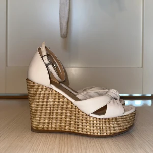 Beige sandaler med flätad platå - Snygga sandaler med hög flätad platå och beige remmar i skinn. Remmarna har en knutdetalj framtill och justerbart spänne vid ankeln. Perfekta för sommarens alla tillfällen och ger extra höjd med sin stabila plattform.