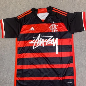 Adidas Flamengo x Stussy fotbollströja L - Säljer en fet fotbollströja från Adidas i samarbete med Flamengo och Stüssy. Tröjan är svart och röd med breda horisontella ränder, vit Stüssy-logga på bröstet och Flamengo-emblem. Klassisk v-ringning.