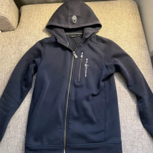 Mörkblå hoodiejacka Sail Racing - Mörkblå hoodiejacka från Sail Racing med dragkedja och bröstficka med logga. Jackan har huva med tryck och långa ärmar. Perfekt för chill dagar och har en clean look med diskreta detaljer.