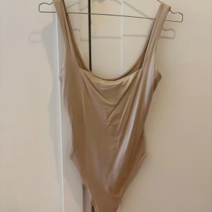 Beige body från Bershka - Den perfekta bodyn från Bershka som snatchar kroppen så snyggt och bekvämt. Fyrkantig, djup urringning både fram och bak. Modellen är enkel och stilren med högt skurna ben (string). Materialet är mjukt och stretchigt syntet som sitter bekvämt mot kroppen. Det perfekta basplagget!! Endast använd en gång. 