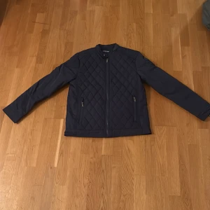 Mörkblå quiltad jacka Hunter Story - Snygg mörkblå quiltad jacka från Hunter Story med dragkedja framtill och två fickor med zip. Jackan har ett stilrent rutmönster och långärmade ärmar med zip-detalj vid mudden. Perfekt för dig som gillar en clean look med lite edge. Den är helt oanvänd.