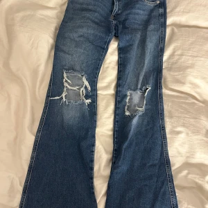 Blå bootcut jeans från 157, L - Snygga blå jeans från 157 i modellen Paris Core, storlek L. Jeansen har bootcut passform och slitna detaljer med stora hål på båda knäna. Tillverkade i klassiskt denimtyg med dragkedja och dubbla knappar i midjan.