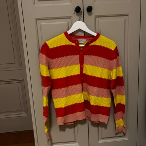 Randig kofta från Stella McCartney - Färgglad kofta från Stella McCartney med breda ränder i rött, gult och rosa. Tröjan har långärmad design, rund hals och knappar framtill. Stickad struktur som ger en cool och unik känsla. Perfekt för dig som gillar att sticka ut med din stil.