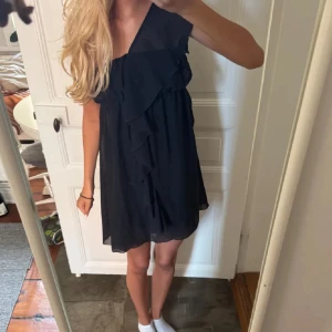 Svart kort volangklänning - Säljer en svart kort klänning med volangdetaljer framtill. Klänningen har en luftig passform och är ärmlös, perfekt för dig som gillar en enkel men snygg stil. Materialet känns lätt och skönt mot huden.