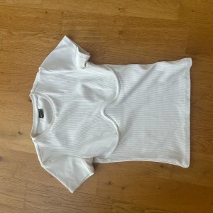Vit ribbad topp från Gina Tricot - Säljer en vit ribbad topp från Gina Tricot i storlek S. Toppen har korta ärmar och en rund halsringning. Den är figurnära med snygga sömdetaljer framtill som ger en cool look. Perfekt att styla med jeans eller kjol för en fräsch vibe.
