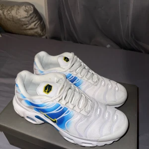Nike Air Max Plus vita/blå sneakers - Nike Air Max Plus sneakers i vitt med blå och ljusblå gradient på sidorna och gula detaljer. Snygg mesh-överdel, synliga Air Max-enheter i sulan och klassisk snörning. Rund tå och platt sula för skön komfort. Perfekt för dig som gillar sportig och modern stil. Pris kan diskuteras