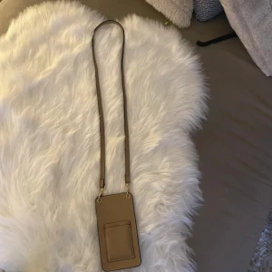 Beige axelväska i skinn - Slimmad beige axelväska med lång justerbar rem och två fack, perfekt för mobilen och kort. Väskan har stilrena detaljer i guld och är tillverkad i slätt skinn. Enkel och snygg design som passar till många outfits.