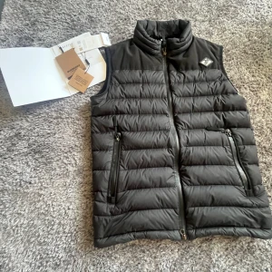 Svart dunväst från Burberry - Svart dunväst från Burberry med quiltad design och hög krage. Västen har två praktiska fickor med dragkedja och en diskret Burberry-logga på bröstet. Materialet är glansigt och känns lyxigt, perfekt för lager-på-lager under kyliga dagar.