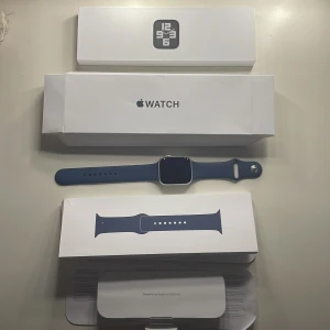 HELT NY. OANVÄND Apple Watch SE 44mm silver/blå - LADDARE MEDFÖLJER+Alla andra tillbehör. Apple Watch SE (Gen 2) med 44mm aluminiumboett i silver och sportarmband i marinblått. Klockan har en stilren, fyrkantig form och digital display. Perfekt för dig som vill ha koll på mobilen direkt på handleden och gillar modern design.