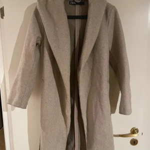 Beige kappa med huva från Zara - Säljer en beige kappa från Zara med huva och bälte i midjan. Jackan har lång ärm, slits i sidorna och en snygg omlott-design framtill. Perfekt för dig som gillar stilrena och enkla plagg med lite extra edge.