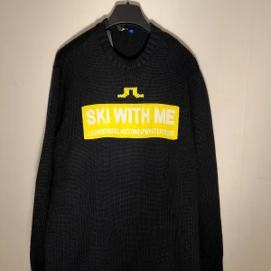 Stickad tröja J.Lindeberg - Svart stickad tröja från J.Lindeberg med gul och vit text 'SKI WITH ME' framtill. Grovstickad modell med rund halsringning och långa ärmar. Perfekt för kalla dagar och snygg detalj med den stora loggan på bröstet.