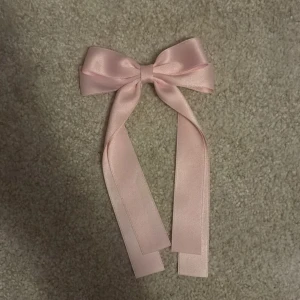 Rosa satinrosett diadem - Söt rosa rosett i satin med långa band och metallspänne på baksidan. Perfekt som håraccessoar för att ge en gullig och trendig touch till din look. Enkel att fästa i håret och ger en mjuk, feminin vibe.