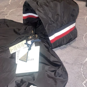 Svart vindjacka från Moncler - Svart vindjacka från Moncler med huva som har snygga ränder i vitt, rött och blått. Jackan är tillverkad i lätt och glansig polyester, har dragkedja framtill och snörning vid huvan. Perfekt för dig som gillar stilrena och sportiga plagg med exklusiv känsla.