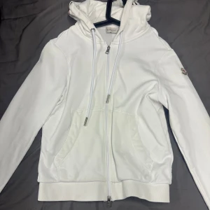 Vit hoodie från Moncler med huva - Vit hoodie från Moncler med dragkedja, huva och snörning. Tröjan har fickor framtill, broderad Moncler-logga på ärmen och ribbade muddar med rödblåa detaljer. Tillverkad i mjuk bomull, perfekt för en clean och sportig look.