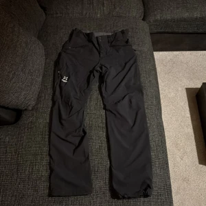 Svarta täckbyxor från Haglöfs - Säljer ett par svarta täckbyxor från Haglöfs med justerbar midja och dragkedjor (dragkedjan på högra fickan är sönder) vid fickorna. Byxorna har en stilren design med logotyp på benet och är gjorda i slitstarkt syntetmaterial som står emot väder och vind. Perfekta för utomhusaktiviteter under vintern.