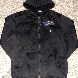 Svart hoodie från Polo Ralph Lauren - Svart hoodie från Polo Ralph Lauren med dragkedja, huva och snörning. Klassisk vit broderad logga på bröstet och två fickor framtill. Perfekt för en chill och stilren look.