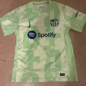 Barcelona fotbollströja Nike grön - Snygg Barcelona fotbollströja från Nike i ljusgrön färg med abstrakt mönster. Tryck med Spotify och FCB-logga på bröstet, samt UNHCR och andra detaljer på ärmarna. Kortärmad modell i lätt och ventilerande material, perfekt för match eller träning.