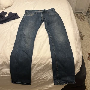 Blå jeans från Lee med straight fit - Säljer ett par klassiska blå jeans från Lee med rak passform och fem fickor. Jeansen har snygga slitningar, kontrastsömmar och dragkedja i gylfen. Perfekt för dig som gillar tidlös stil och vill ha ett par jeans som funkar till allt.