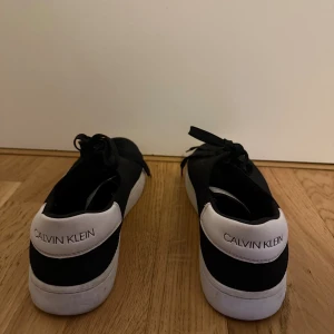 Svarta sneakers från Calvin Klein - Svarta sneakers från Calvin Klein med vit häl och vit platt sula. Skorna har snörning och är tillverkade i ett syntetmaterial med meshstruktur på ovansidan. Calvin Klein-logga syns tydligt på hälen och sidan av sulan. Perfekt för dig som gillar stilrena och klassiska sneakers.