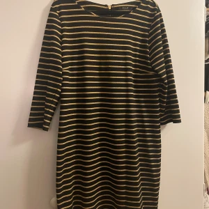 Snygg svart och guld randig klänning, Vila - Svart kortklänning från Vila med horisontella guldiga ränder. Klänningen har trekvartsärm, rund halsringning och dragkedja bak. Materialet är mjukt och stretchigt, vilket gör den bekväm och enkel att bära. Perfekt för dig som gillar en stilren look med lite extra glans. Bra skick! 