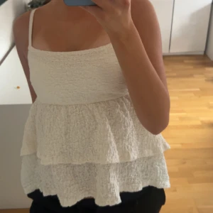Vit volang linnetopp från Gina Tricot - Supersöt vit linnetopp från Gina Tricot i storlek S. Toppen har smala axelband, volang i två lager och ett krusat, texturerat tyg som ger en cool och luftig känsla. Perfekt för varma dagar och enkel att styla med jeans eller kjol.