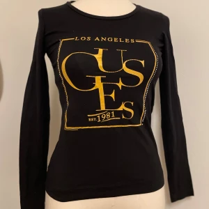 Svart långärmad tröja från Guess - Svart långärmad tröja från Guess med stort gult tryck på bröstet där det står 'Los Angeles' och 'Est. 1981'. Tröjan har en rund halsringning och är figurnära i modellen. Perfekt för dig som gillar statement-plagg och snygga loggor.