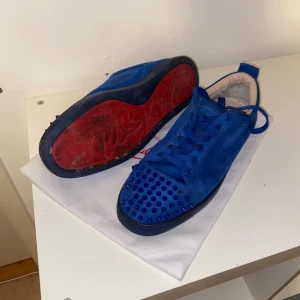 Blå Christian Louboutin sneakers - Säljer ett par blå sneakers från Christian Louboutin med coola nitar på tån och klassisk röd sula. Skorna har snörning och är gjorda i mocka med en rund tå. Perfekt för dig som vill sticka ut med lyxig streetstyle. Några nitar har lossnat det är bara att köpa nitar🤩 hör av er för fler frågor lite smutsiga 