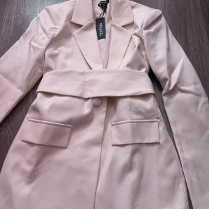 Krämvit kavaj med bälte från Boohoo - Snygg krämvit kavaj från Boohoo med markerad midja och tillhörande bälte. Kavajen har klassisk krage, en dold knapp framtill och två fickor med lock. Materialet är slätt och har en lätt glansig finish som ger en exklusiv känsla. Kavajen är helt oanvänd med prislapp på!