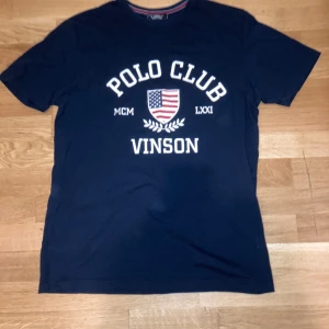 Mörkblå Vinson Polo Club T-Shirt - Snygg mörkblå t-shirt från Vinson Polo Club med vit tryckt text och amerikanskt sköldmotiv på bröstet. Rund hals och klassisk passform. Perfekt för dig som gillar sportig och stilren design. Materialet är mjuk bomull.
