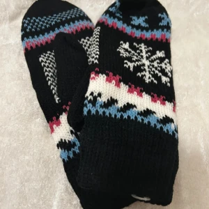 Svarta stickade vantar med snöflinga - Mysiga svarta stickade vantar med klassiskt vintermönster i vitt, blått och rött. Ena vanten har en stor snöflinga på ovansidan och båda har flera ränder och mönsterdetaljer. Perfekta för kalla vinterdagar.