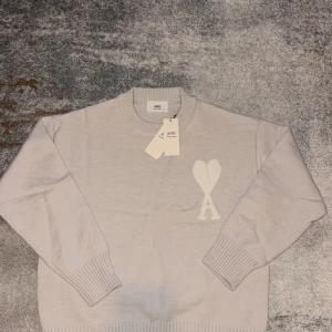 Ljusbeige stickad tröja från Ami - Säljer en ljusbeige stickad tröja från Ami med rund hals och ribbade muddar. På bröstet finns ett stort vitt Ami-hjärta med A. Tröjan har lång ärm och en loose passform, perfekt för dig som gillar stilren design.