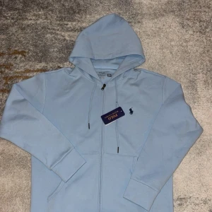 Ljusblå hoodie från Polo Ralph Lauren - Snygg ljusblå hoodie från Polo Ralph Lauren med dragkedja och huva. Tröjan har snörning vid halsen, ribbade muddar och det klassiska Polo-logot på bröstet. Perfekt för chill dagar och enkel att matcha med jeans eller joggers.