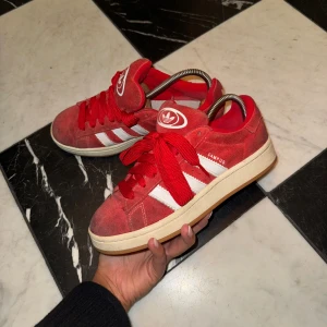Röda Adidas Campus sneakers 39 - Säljer ett par röda Adidas Campus sneakers med klassiska vita ränder på sidorna och vit logga på plösen. Skorna har snörning, rund tå och platt sula. Ovandelen är i mocka och insidan är mjukt fodrad. Perfekt för dig som gillar retrostil och streetwear.