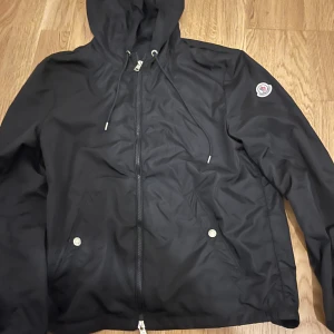 Svart vindjacka från Moncler - Svart vindjacka från Moncler med huva och dragsko, snygga silverfärgade knappar på fickorna och klassisk Moncler-logga på ärmen. Jackan har hel dragkedja framtill och är tillverkad i polyester, perfekt för dig som gillar stilren och sportig look.
