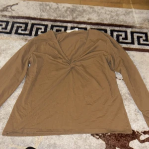 Brun långärmad topp med v-ringning - Säljer en brun långärmad topp med snygg v-ringning och twistad detalj framtill. Toppen har en enkel och clean design som är lätt att matcha med olika outfits. Materialet känns mjukt och bekvämt mot huden, perfekt för dig som gillar stilrena plagg.