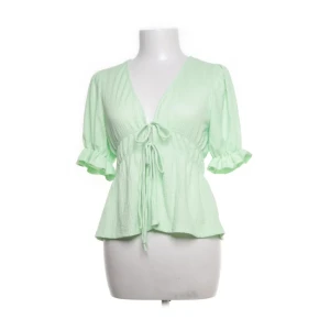 Mintgrön blus med knyt från Gina Tricot - Använd endast en gång. Superfin mintgrön blus från Gina Tricot i ett krispigt, strukturerat polyestertyg. Blusen har v-ringning, korta puffärmar med volangkant och snygg knytning framtill som ger en feminin siluett. Perfekt för dig som gillar trendiga detaljer och pastellfärger. 