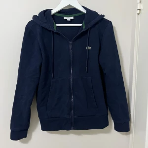 Marinblå hoodie från Lacoste - Snygg marinblå hoodie från Lacoste med klassisk krokodillogga på bröstet. Tröjan har huva med snörning, dragkedja framtill och två stora fickor. Tillverkad i mjuk bomull och har långärmad design med ribbade muddar. Perfekt för chill dagar.