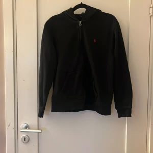 Svart hoodie från Polo Ralph Lauren - Svart hoodie från Polo Ralph Lauren med dragkedja framtill och klassisk röd logga på bröstet. Tröjan har huva, känguruficka och långa ärmar. Perfekt för en chill och stilren look.