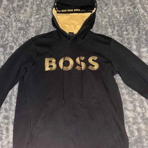Svart hoodie från BOSS med guldtryck - Svart hoodie från BOSS med stort guldigt BOSS-tryck på bröstet och gul insida i huvan. Klassisk känguruficka framtill, snörning i huvan och BOSS-logga längs kanten på insidan av huvan. Perfekt för en chill och snygg streetstil.