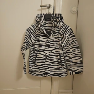Zebramönstrad regnjacka från Funk - Snygg regnjacka från Funk i storlek 140 med svartvita zebraränder. Jackan har huva, dragkedja och två fickor med dragkedjor. Perfekt för dig som vill sticka ut med ett coolt mönster även när det regnar.