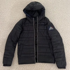 Svart pufferjacka från Scotch & Soda - Svart pufferjacka från Scotch & Soda med huva. Snygga mörkblå och vita detaljer. Bra jacka och mycket bra skick. NYPRIS: 2500