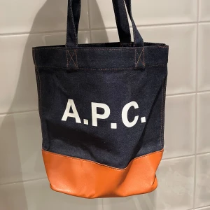 A.P.C. shoppingväska i denim och skinn - Snygg tote bag från A.P.C. i mörkblå denim med orange skinnbotten. Väskan har vita tryckta bokstäver på framsidan och praktiskt innerfack med dragkedja. Perfekt storlek för att bära med sig allt man behöver och cool kontrast mellan material och färger.