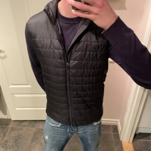 Svart dunväst från On Our Terms - Svart quiltad dunväst från On Our Terms med ståkrage och dragkedja framtill. Västen har två sidofickor med dragkedja och en stilren, sportig look. Perfekt att slänga över en hoodie eller tröja när det är lite kyligt ute.