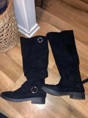 Svarta höga mockastövlar med ringdetalj - Säljer ett par svarta höga stövlar i mjuk mocka med rund tå och platt sula. Stövlarna har snygga guldfärgade ringdetaljer upptill och vid ankeln. Perfekta till höst- och vinterlooken och passar till både jeans och kjol.