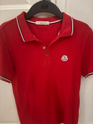 Röd pikétröja från Moncler - Snygg röd pikétröja från Moncler med klassisk krage och två knappar framtill. Tröjan har vita och marinblå ränder på krage och ärmslut samt Moncler-logga broderad på bröstet. Perfekt för dig som gillar stilrena och sportiga plagg.