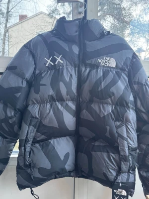 Svart camo pufferjacka från The North Face - Säljer en fet pufferjacka från The North Face x KAWS med svart och grått camo-mönster. Jackan har broderad logga, dragkedja framtill och två sidofickor. Puffig och varm design, perfekt för kalla dagar. Stilsäker look med unika XX-detaljer på bröstet.storlek är XL men passar mer som L 