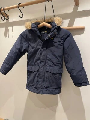 Mörkblå parkajacka från Lyle & Scott - Parkajacka från Lyle & Scott i mörkblått med fuskpäls på huvan och klassisk gul logga på bröstet. Jackan har stora fickor framtill, dragkedja och knappar. Hör av er om ni har några funderingar eller frågor. 