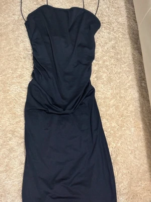 Svart långklänning med smala axelband - Snygg svart långklänning med smala axelband och draperad detalj framtill. Klänningen har en stilren och enkel design som ger en elegant siluett. Materialet känns mjukt och har en lätt glans, vilket gör den extra fin. Perfekt för dig som gillar minimalistisk stil.