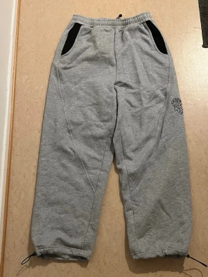 Grå mjukisbyxor från Merta - Säljer ett par grå mjukisbyxor från Merta med svart tryck på benet och bakfickan. Byxorna har svarta detaljer vid fickorna, elastisk midja med snörning och dragsko vid bensluten. Oanvänd, perfekt skick.